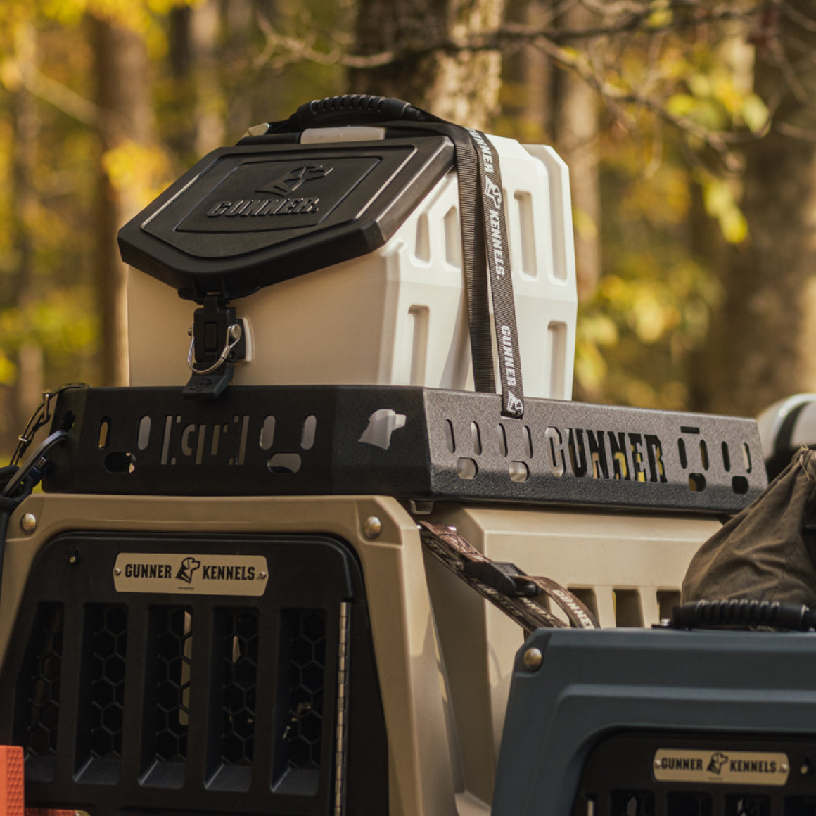 A white Gunner Kennels dog crate and a black Gunner-branded container are secured to the back of a vehicle in a wooded outdoor setting, ready to safely transport your hunting dog through the trees.