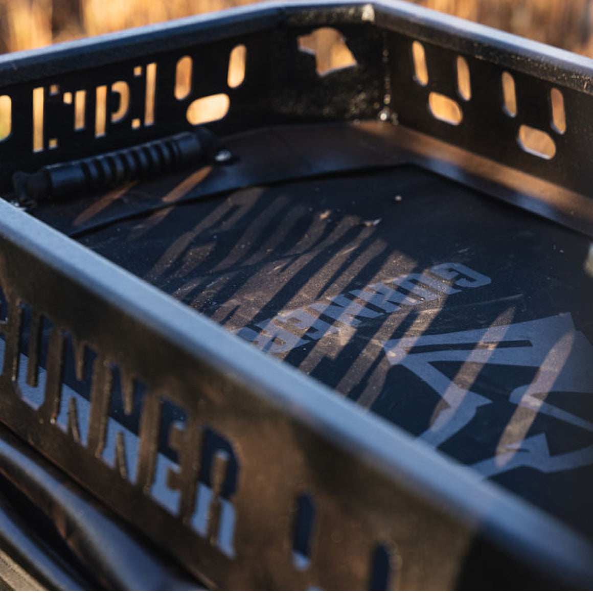 Close-up of a black metal roof rack basket with cut-out letters and angled bars, sunlight casting patterns and shadows on the surface. A kennel handle or rod is visible inside the basket.