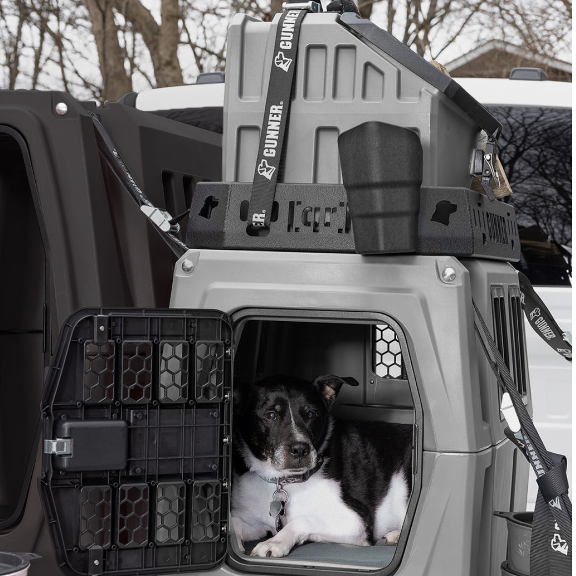 A black and white working dog lies inside a sturdy gray dog crate with an open metal door, while another crate is stacked on top, secured with straps. The crates are in the back of a vehicle outdoors.