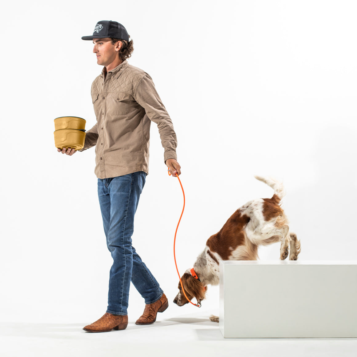 A man in a cap, brown shirt, jeans, and cowboy boots walks a brown-and-white dog on an orange leash at Color Fetch 2025; as the dog climbs onto a white platform, he holds two GUNNER DOG BOWL - Marigold bowls from GUNNER.