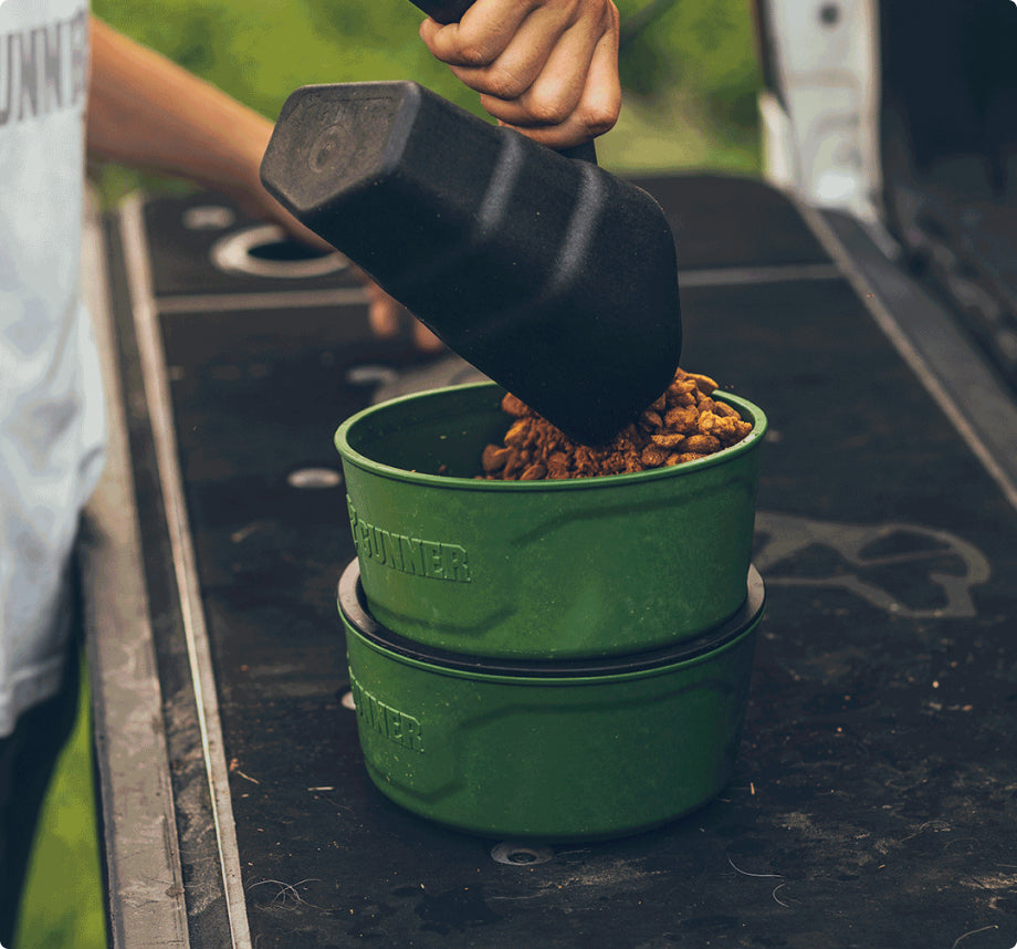 A person scoops brown pet food into a green bowl stacked on another, set on a black outdoor surface—an ideal setup near your dog kennel or crate for organized outdoor gear.