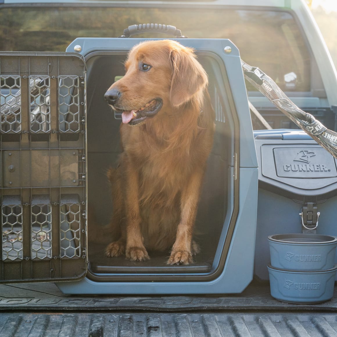 A golden retriever sits inside an open Ducks Unlimited Blue G1™ Kennel by GUNNER on a pickup truck bed, with sunlight in the background and two stacked water bowls—ideal for waterfowl trips or Ducks Unlimited events.