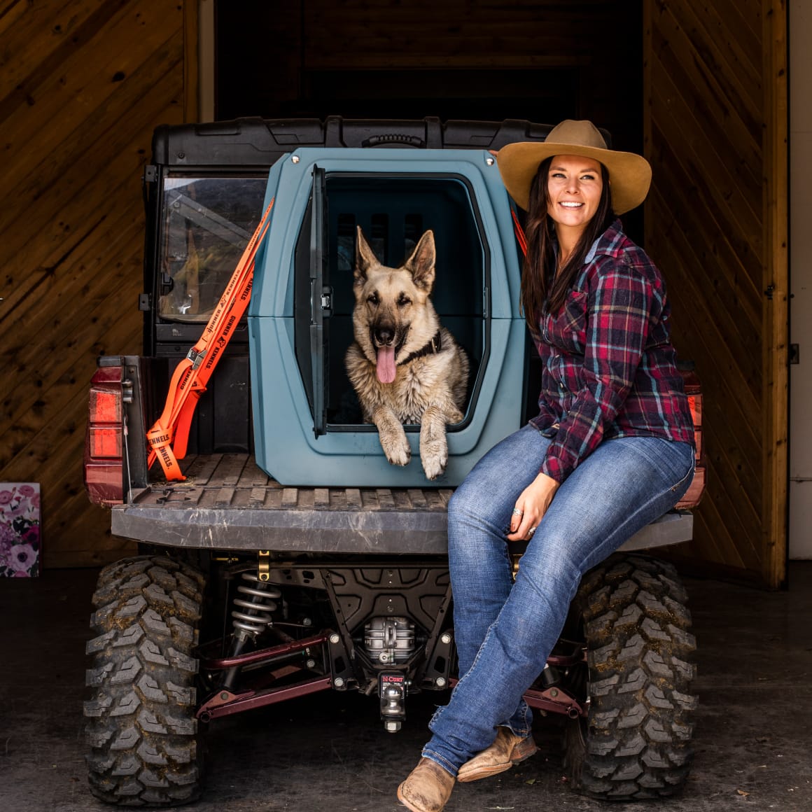 A woman in a hat and plaid shirt smiles beside her German Shepherd in a GUNNER G1™ KENNEL - Ducks Unlimited Blue on an off-road vehicle, both happy after exploring waterfowl habitats with Ducks Unlimited.