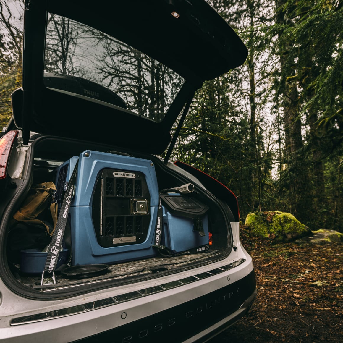 An open SUV trunk in a forest holds the GUNNER G1™ KENNEL - Ducks Unlimited Blue and camping gear, ideal for exploring waterfowl habitats, with trees and mossy rocks in the background.