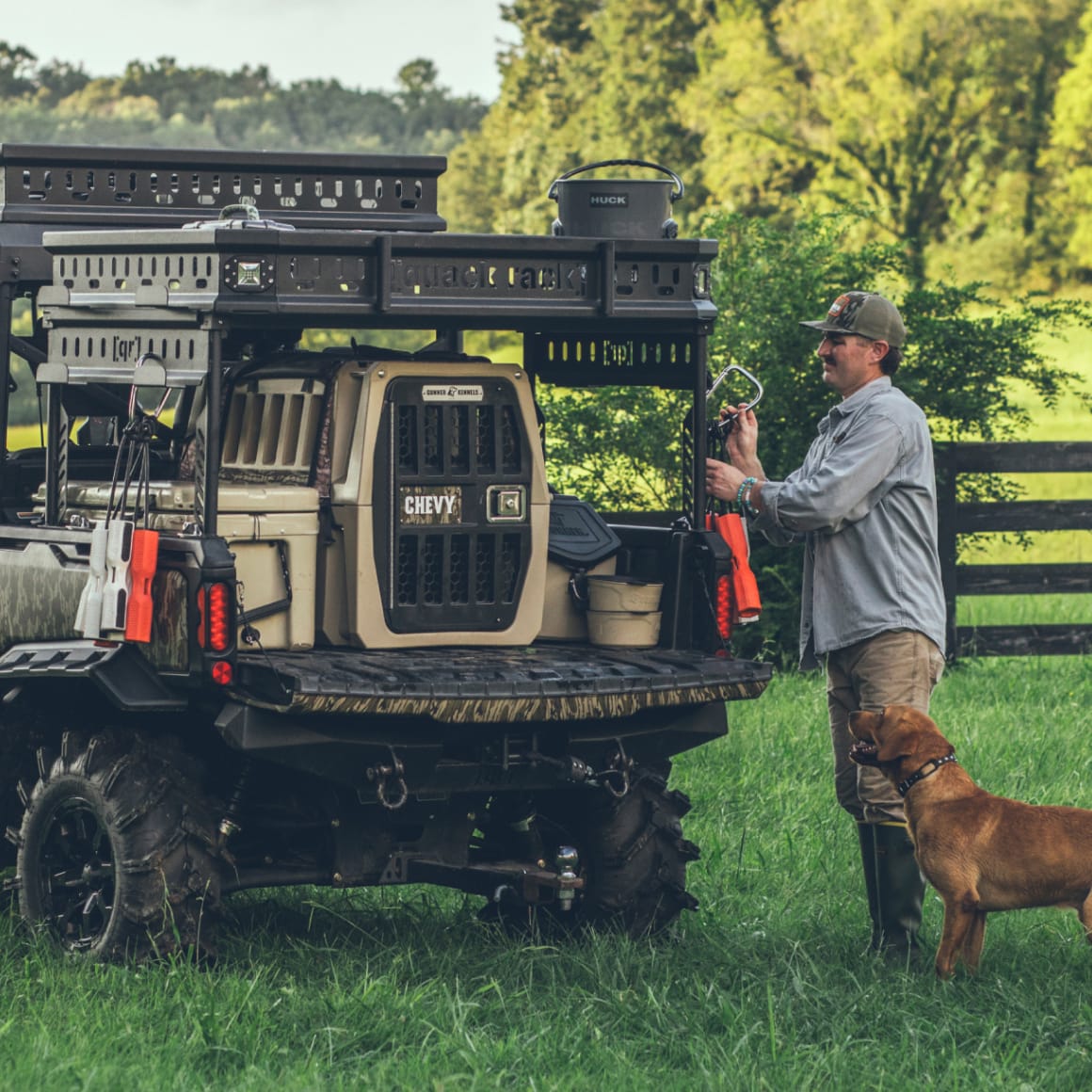 A man in outdoor gear stands by an off-road vehicle equipped with the GUNNER G1™ KENNEL - TAN for shock protection, supplies in the back, and a brown dog nearby in a grassy field with trees and a wooden fence.