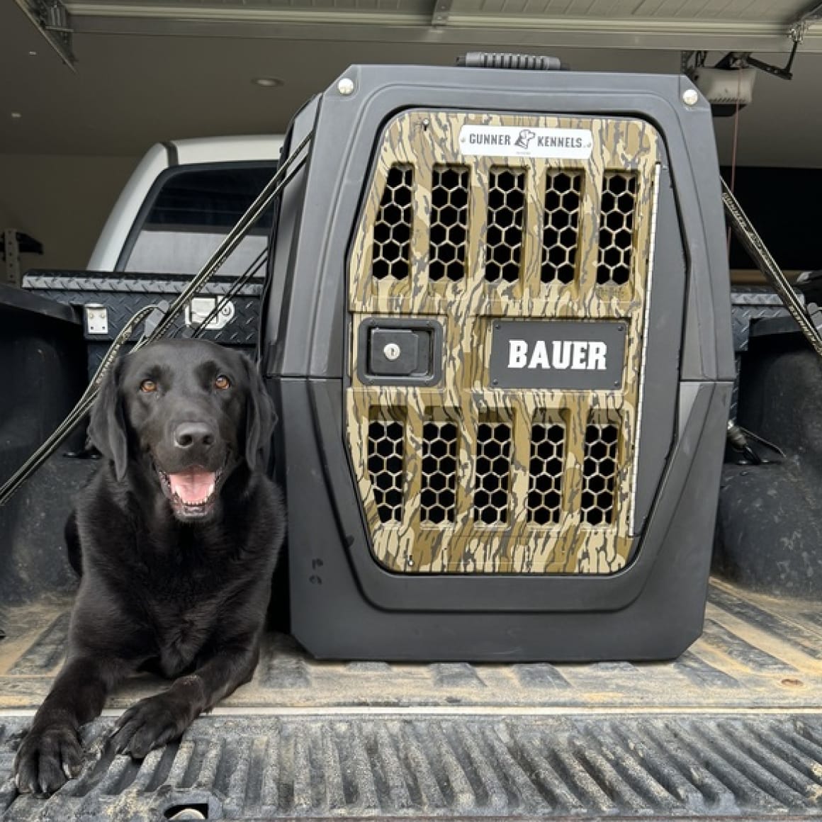 A black labrador rests beside a GUNNER G1™ KENNEL - MOSSY OAK BOTTOMLAND BLACK in the back of a truck parked in a garage.