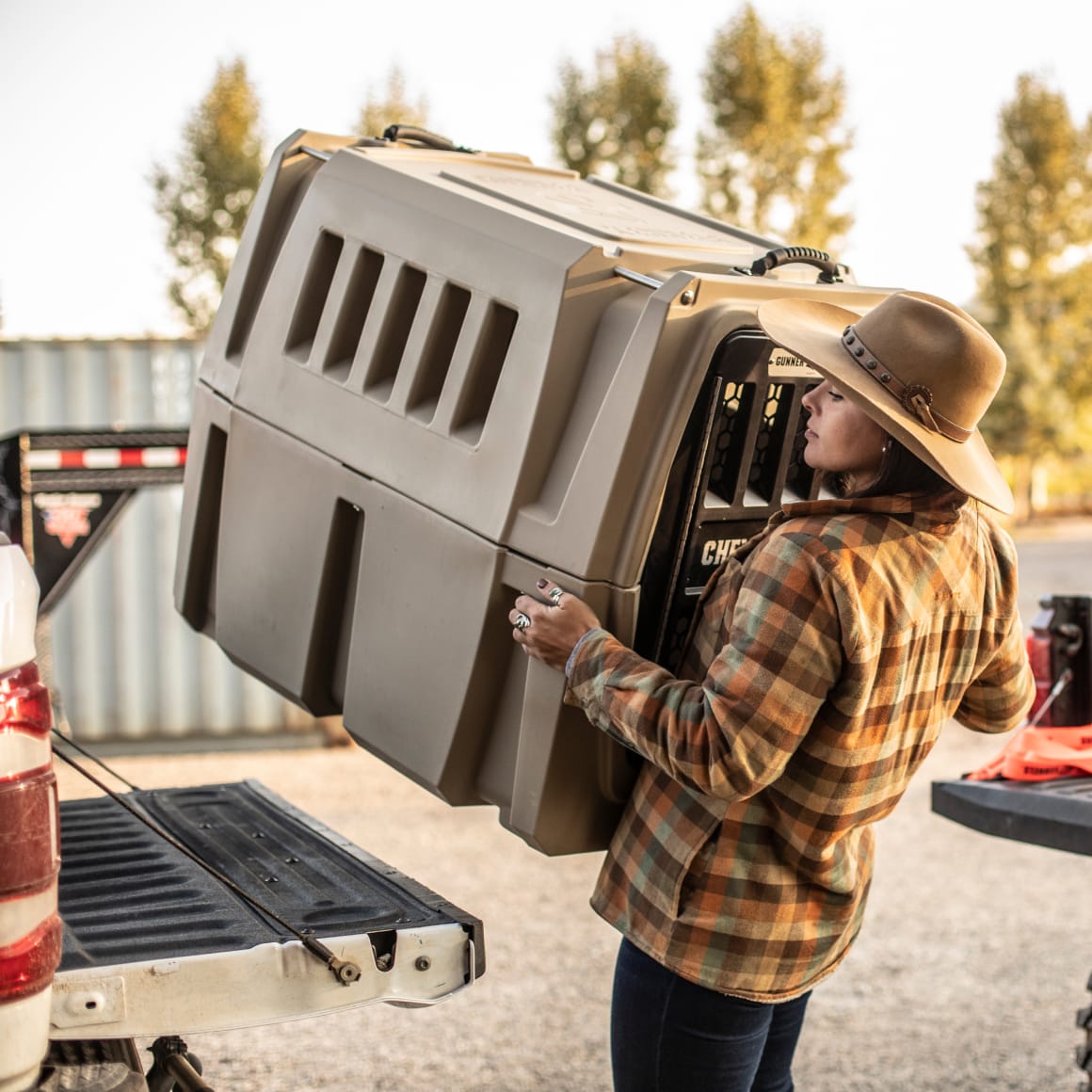 Wearing a plaid shirt and wide-brim hat, a person lifts the GUNNER G1™ KENNEL - TAN from their pickup truck bed outdoors, with trees and a metal storage container behind them, ensuring shock protection for their pet.