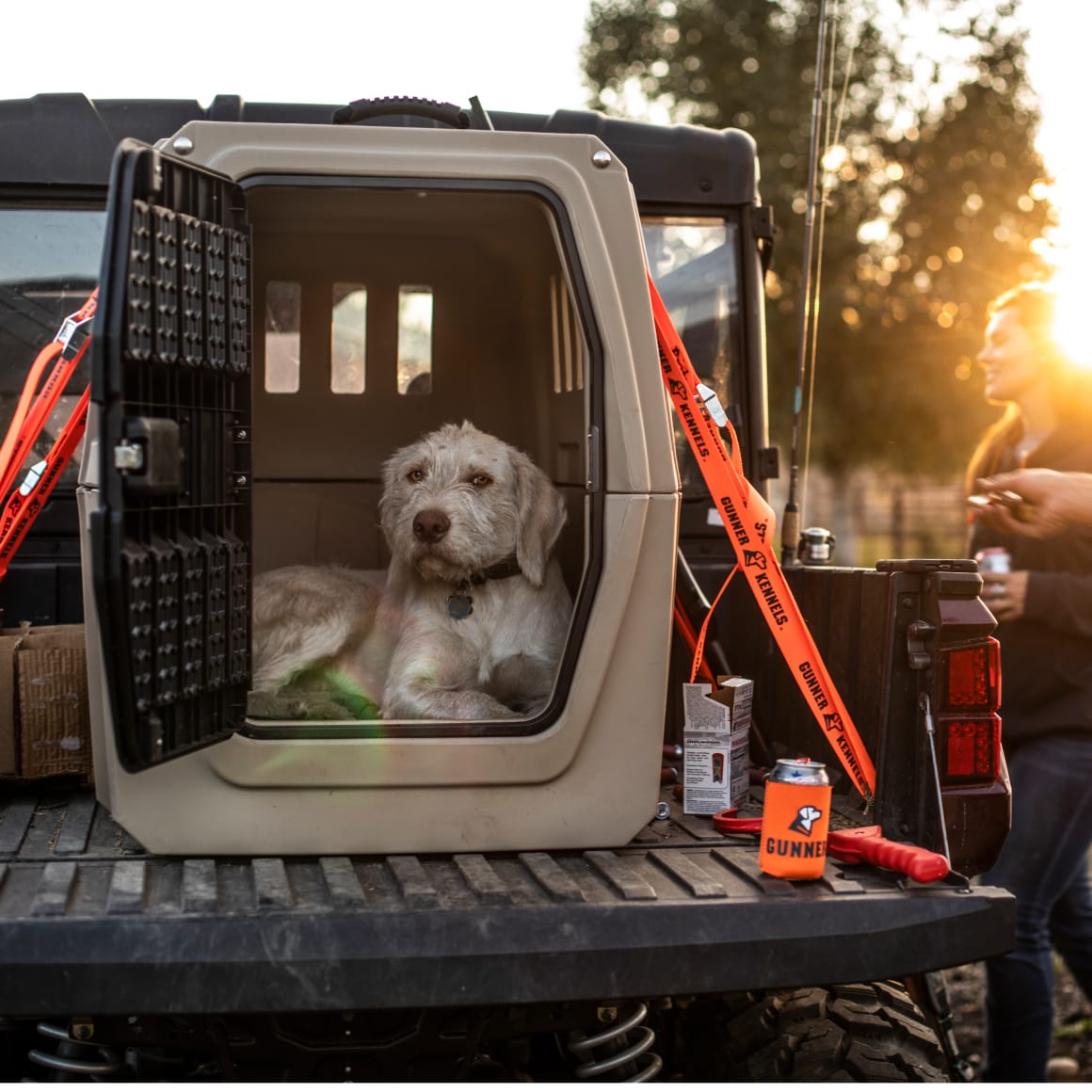 A light-colored dog relaxes in the GUNNER G1™ KENNEL - MOSSY OAK BOTTOMLAND TAN on a vehicle’s tailgate at sunset. An orange leash hangs from the kennel as a woman stands nearby, with trees in the background.