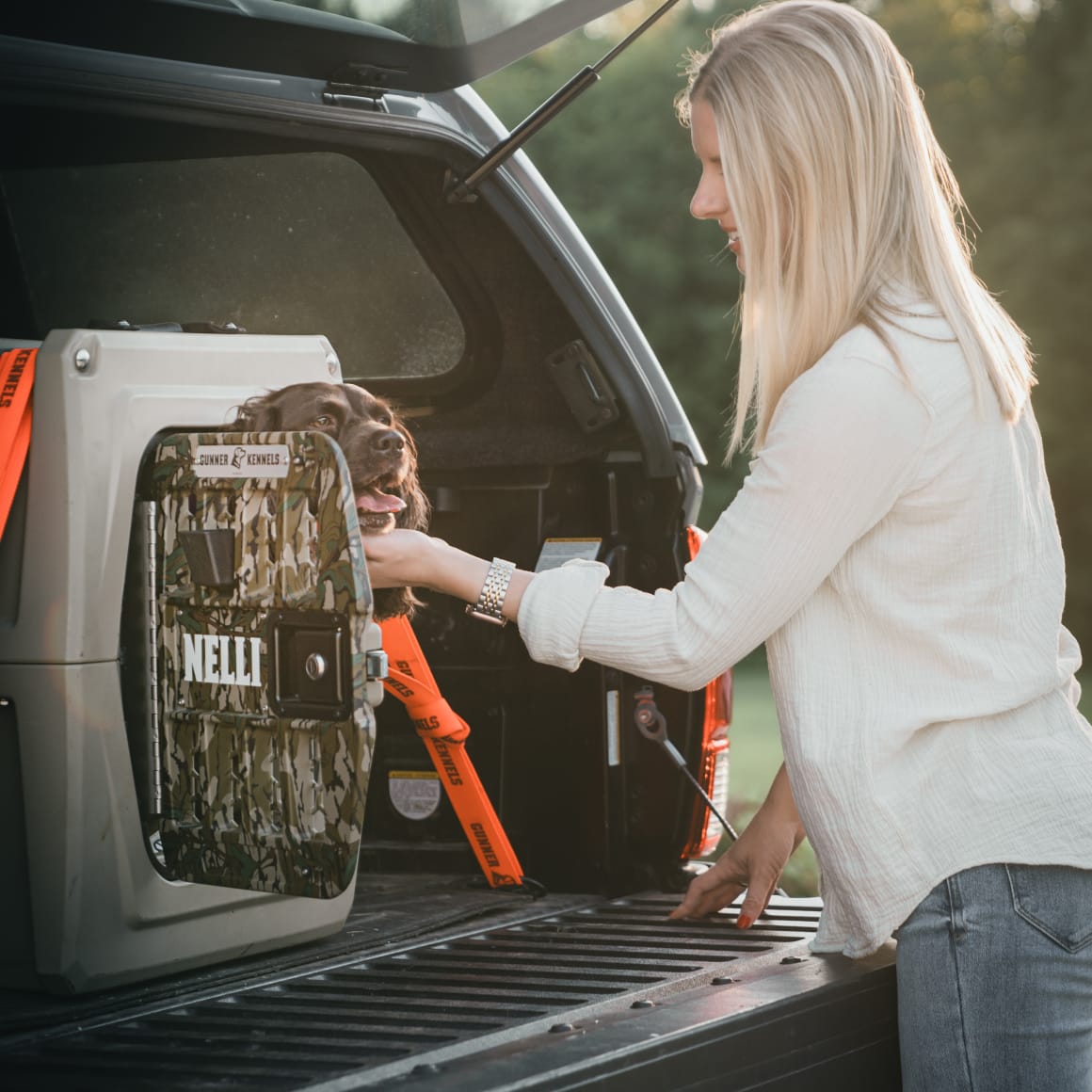 A woman with long blond hair smiles and pets a brown dog named Nelli inside a GUNNER G1™ KENNEL - MOSSY OAK BOTTOMLAND TAN, camouflaged in the back of a pickup truck with the tailgate down.