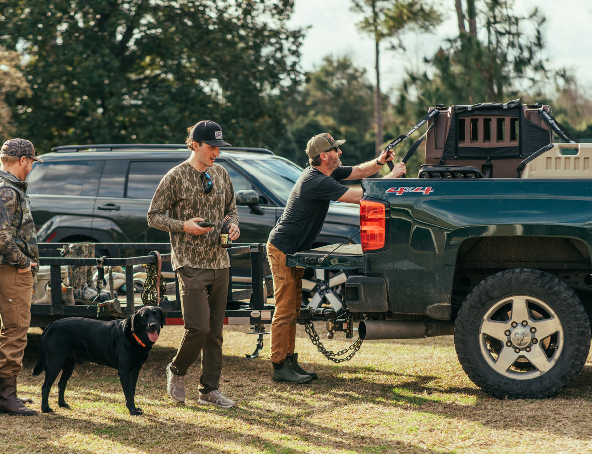 Three men and a black dog stand by a green pickup, as one loads gear into the bed near their GUNNER G1™ KENNEL - SUSQUEHANNA. The scene captures a classic moment for the GUNNER Archive Series, with trees and another vehicle in the background.