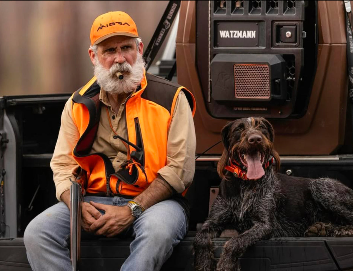 An older man with a cigar and rifle sits beside a happy hunting dog on the back of a vehicle, showcasing the GUNNER G1™ KENNEL - SUSQUEHANNA to help protect dogs on every outdoor adventure.