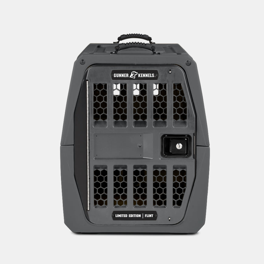 A gray, hard-sided hunting dog kennel with a secure door featuring honeycomb vents, a handle on top, and Gunner Kennels and Limited Edition | Flint labels on the front.