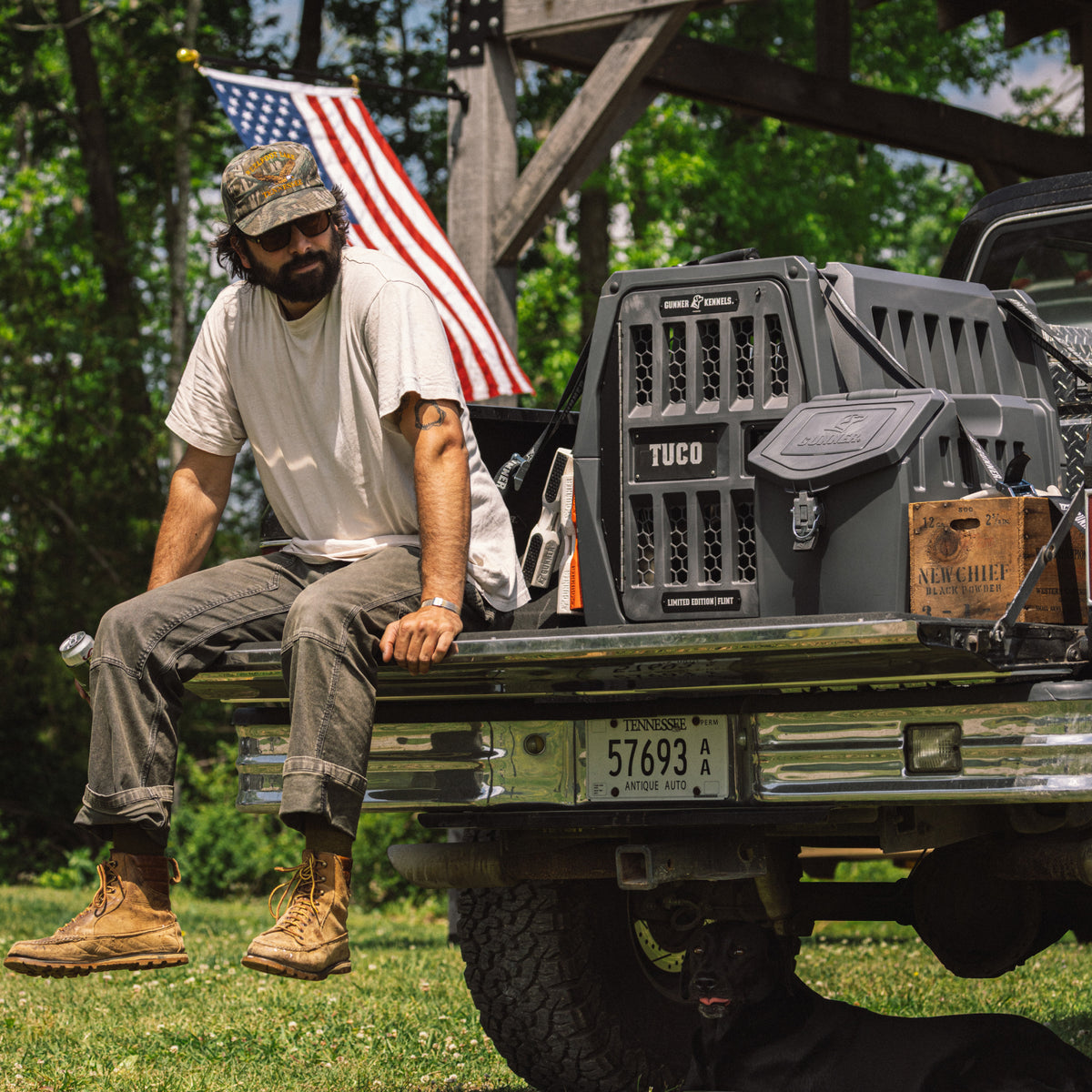 A man in a camo hat, sunglasses, and work boots sits on a rugged pickup next to the GUNNER G1™ KENNEL - FLINT. An American flag hangs nearby while Flint, a black dog, relaxes under the truck among tall trees.