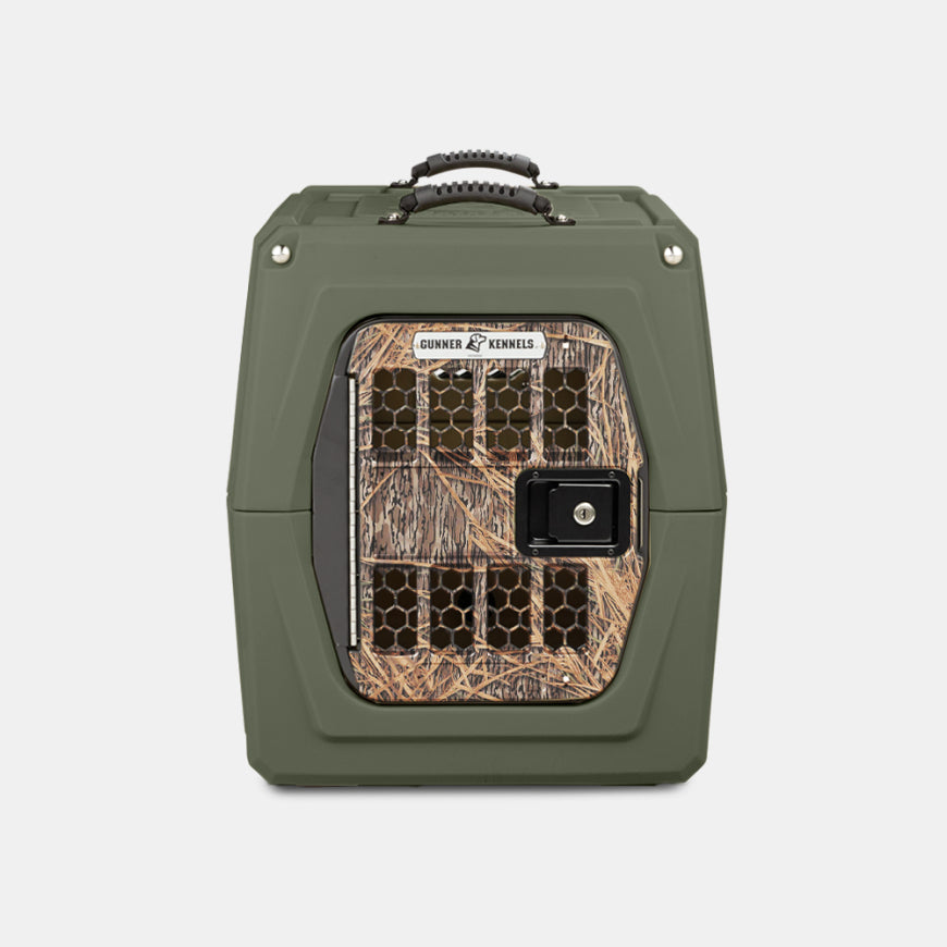 A green, hard-sided pet crate designed for hunting dogs, with a handle on top and a front door featuring a camouflage pattern and ventilation holes, perfect as outdoor gear against a plain white background.