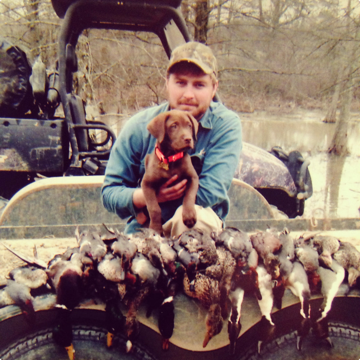 A man in camouflage holds a brown hunting dog puppy while sitting on a boat. The boats edge is lined with several ducks, and a muddy forested area is visible in the background.