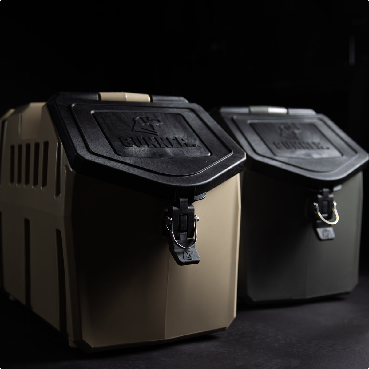 Two sturdy, plastic dog kennel storage containers with black lids and metal latches sit side by side on a dark surface—one tan, one olive green. Both lids are embossed with the GUNNER logo, perfect for the working dogs gear.