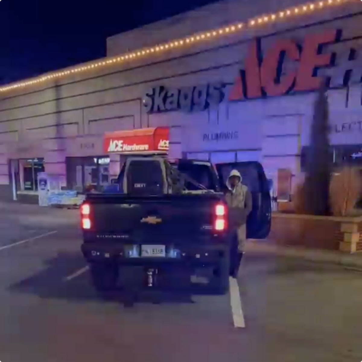 A person stands next to an open door of a black pickup truck parked outside an Ace Hardware store at night, illuminated by bright lights, with outdoor gear visible in the truck bed.