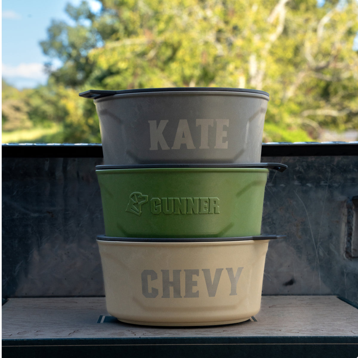Three pet bowls stacked on top of each other—perfect for any kennel—labeled KATE (gray), GUNNER (green with a paw print), and CHEVY (beige), with trees in the background. Ideal for your hardworking hunting or working dog.