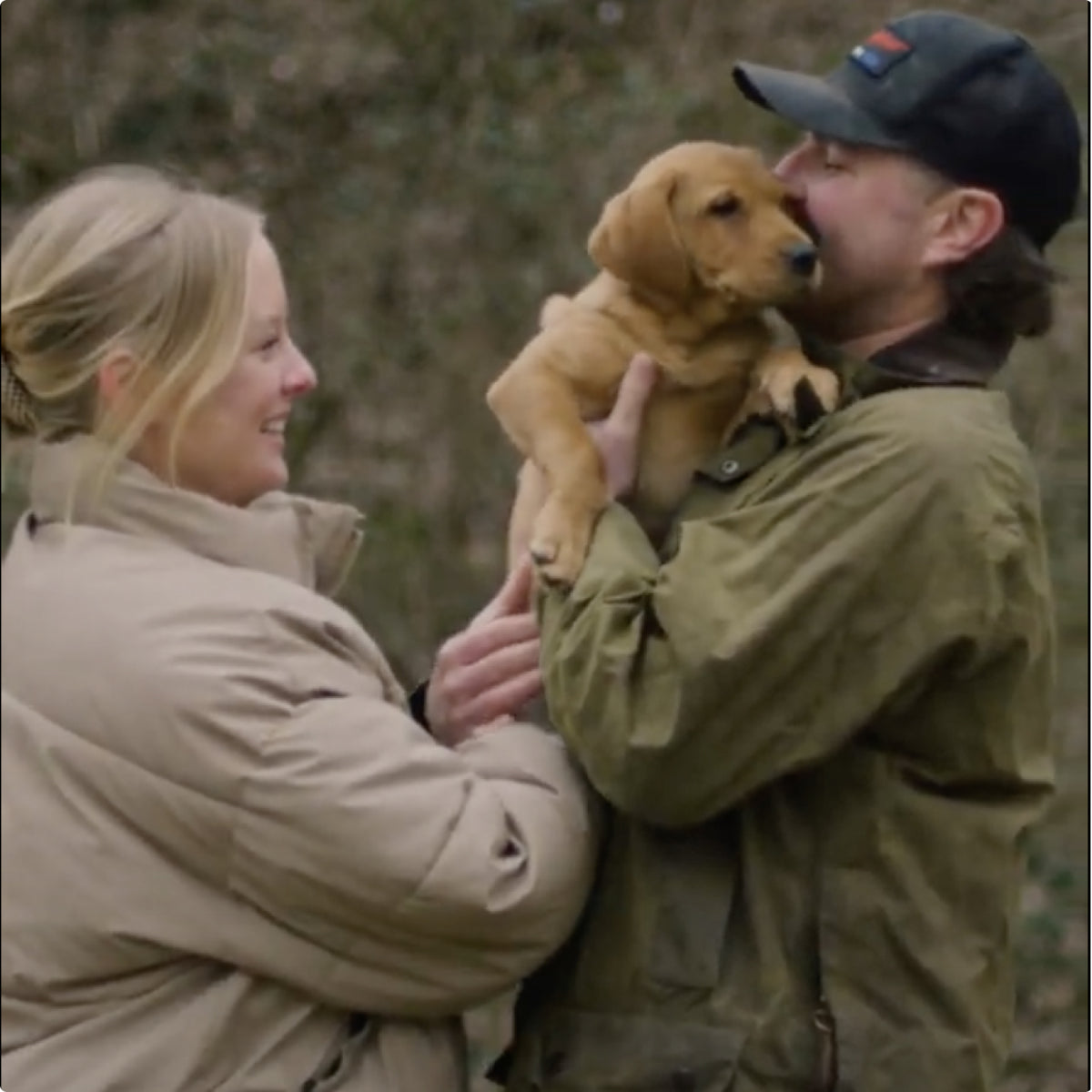 A smiling woman and man stand outdoors, both wearing jackets. The man holds a light brown hunting dog puppy up to his face, while the woman reaches out to pet the puppy.