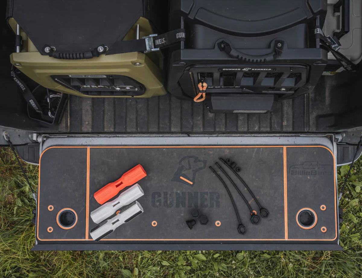 A truck tailgate with a GUNNER-branded mat displays GUNNER BUMPERS, rubber straps, and fasteners for training. Two large dog crates sit in the truck bed above the tailgate. Grass surrounds the parked truck.