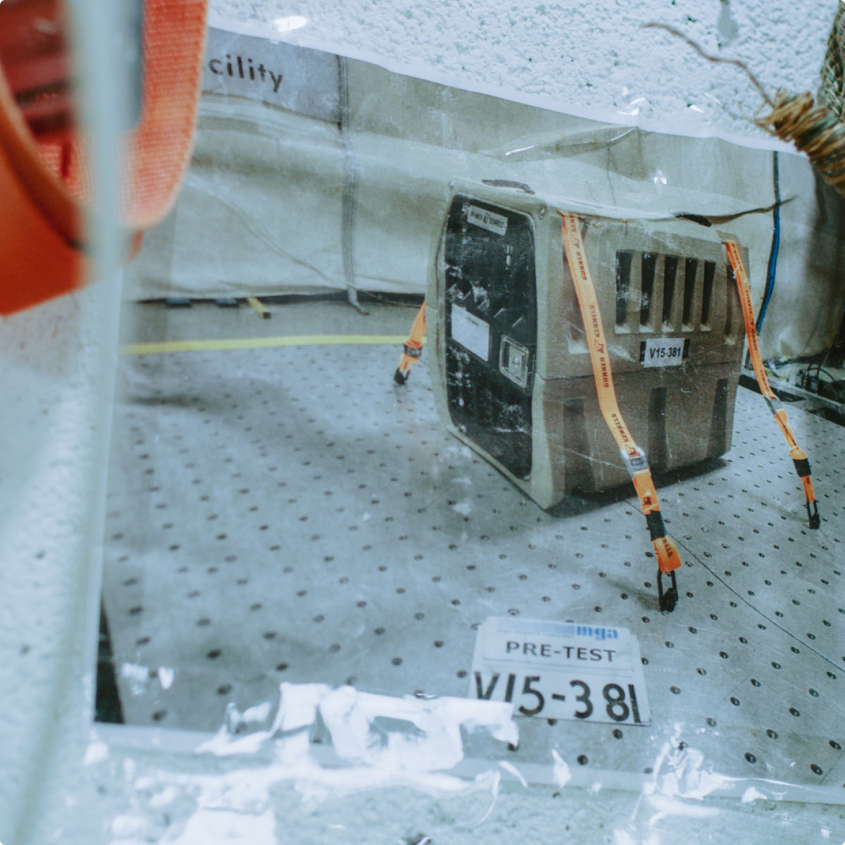 A photograph of a dog kennel secured with orange straps to a perforated metal floor, with a PRE-TEST sign labeled V15-381 placed in front of it at a testing facility.