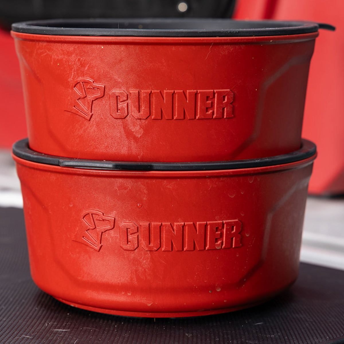 Two stacked red GUNNER DOG BOWLs by GUNNER, each embossed with GUNNER and a dog head logo, ideal for travel, set against a blurred background.