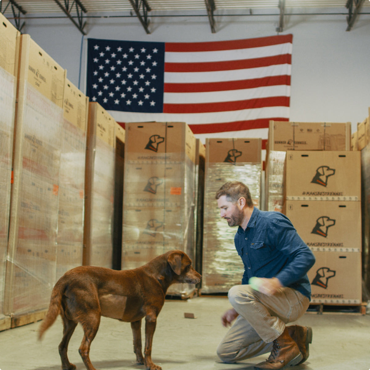 A man kneels on the floor of a warehouse filled with stacked boxes, facing his loyal working dog. An American flag hangs on the wall behind them, hinting at their shared dedication to outdoor gear and adventure.