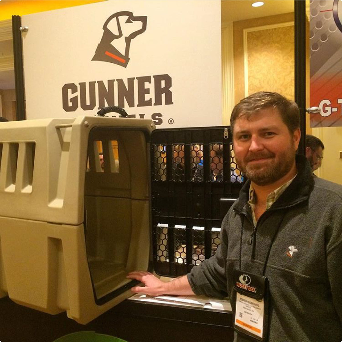 A man with a beard stands next to a large beige dog crate at a booth display for Gunner Kennels, featuring a dog logo and branded signage in the background.