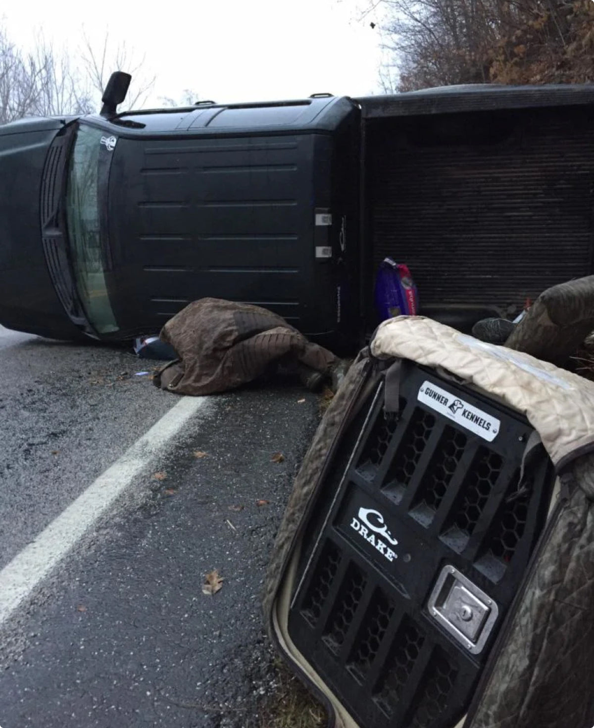 A black pickup truck lies on its side off a wet, rural road. A covered figure is motionless nearby. A large dog crate sits in the foreground, with trees and an overcast sky in the background.