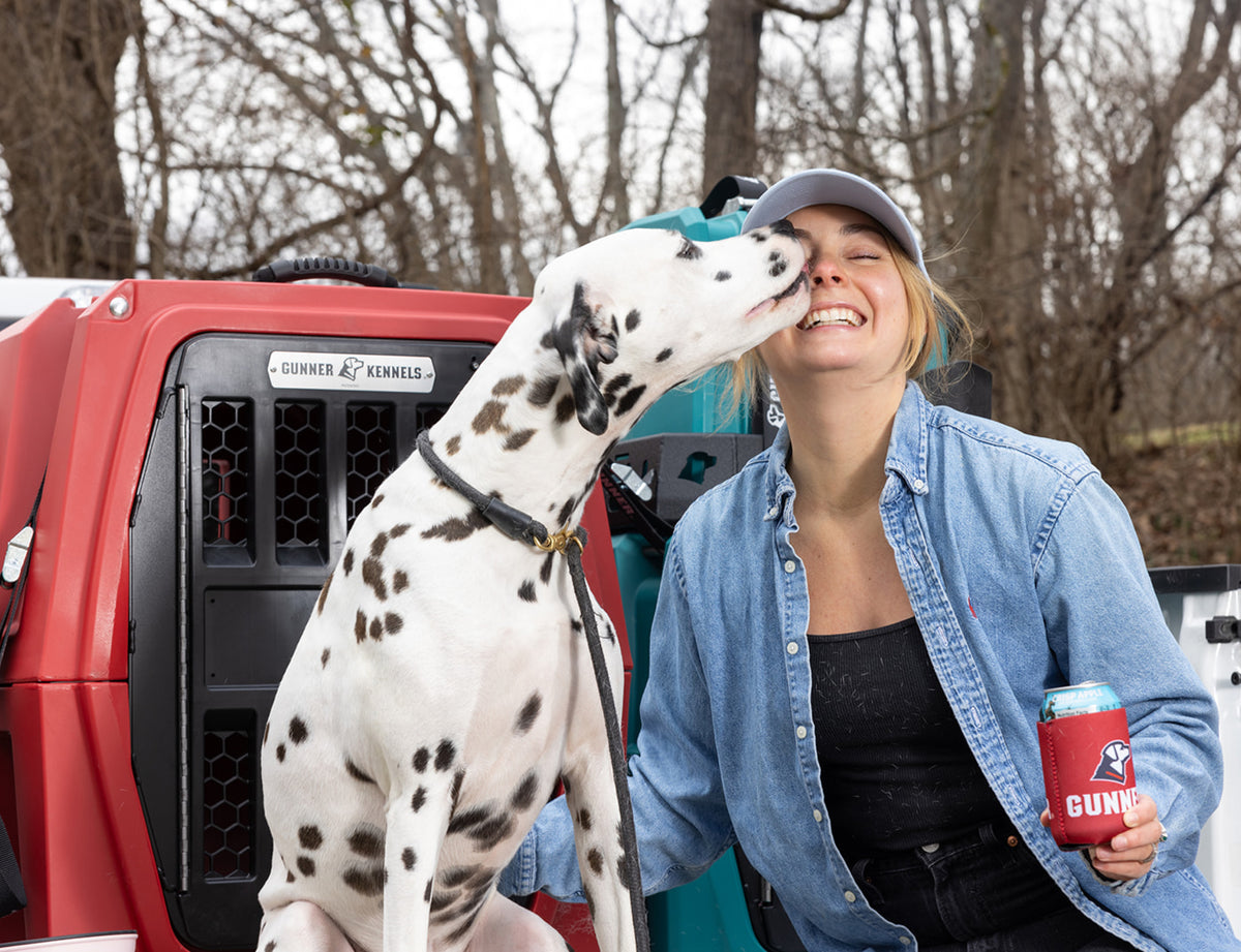 A smiling woman in a denim shirt holds a drink and gets licked on the face by a Dalmatian hunting dog in front of a red pet crate, outdoors with trees in the background.