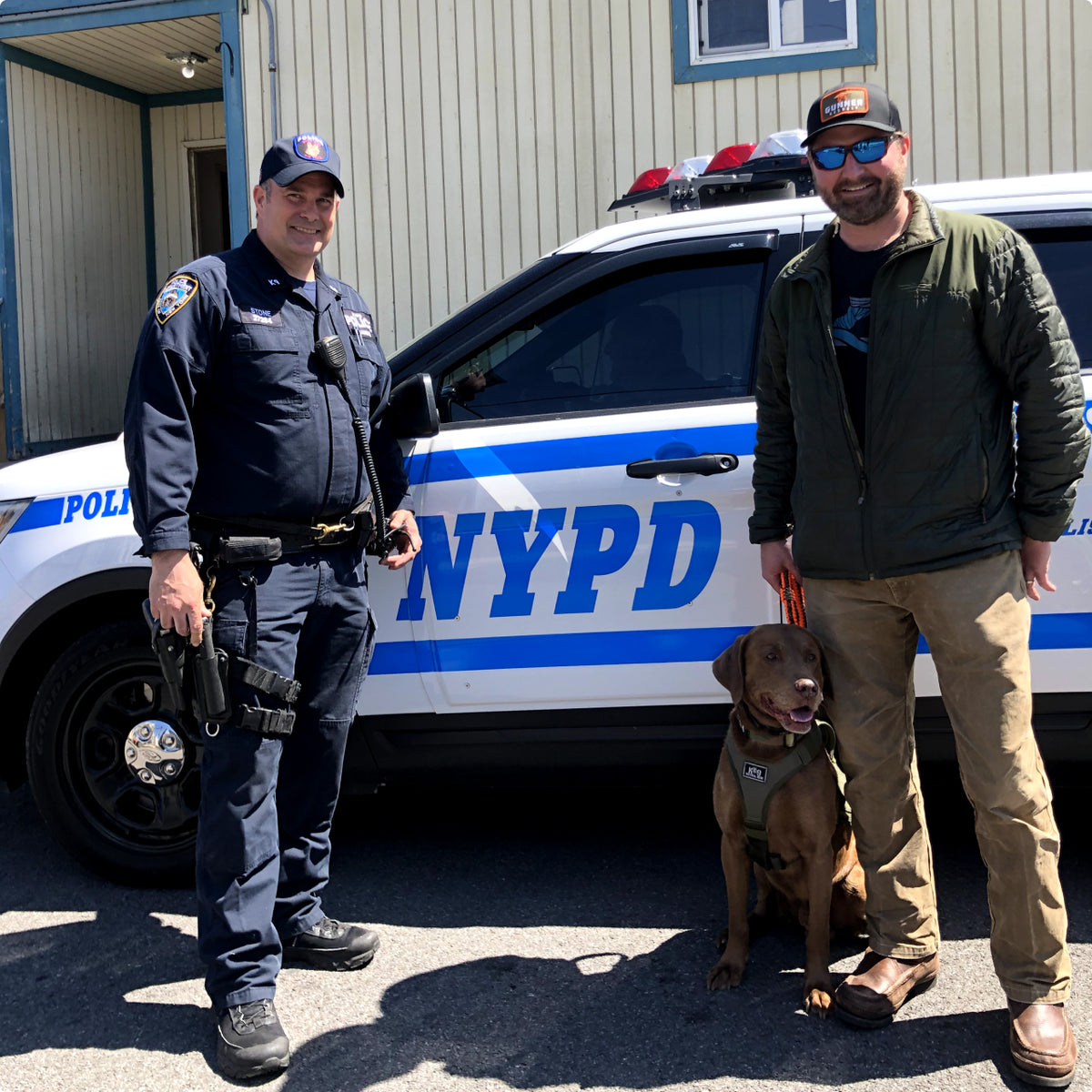 A police officer stands beside a man and a brown hunting dog in front of an NYPD patrol car parked on the street. The officer is in uniform, while the man sports sunglasses, a cap, and outdoor gear.