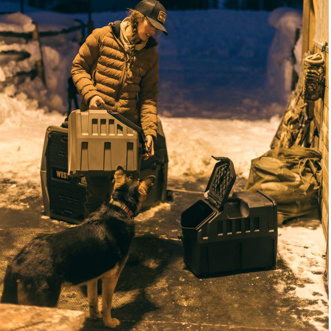 A person in a brown jacket and cap prepares a GUNNER FOOD CRATE - Black by GUNNER outside in the snow, while a German Shepherd stands nearby watching.