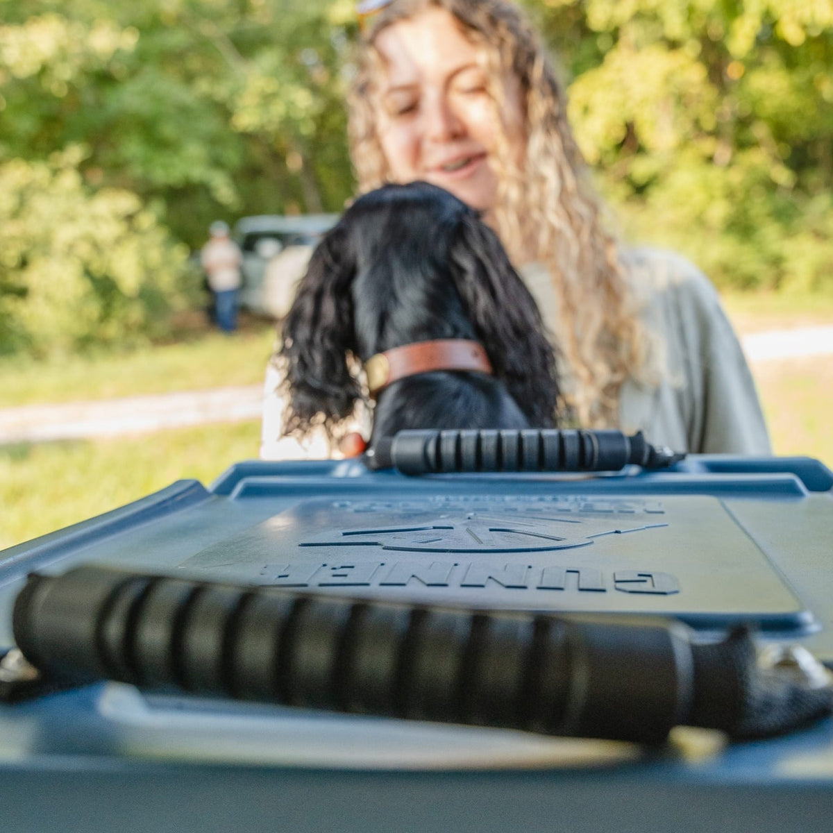 A woman with curly hair smiles at a black dog wearing a brown collar, standing behind the GUNNER G1™ KENNEL - Ducks Unlimited Blue, outdoors with trees and grass—ideal for waterfowl habitat adventures.