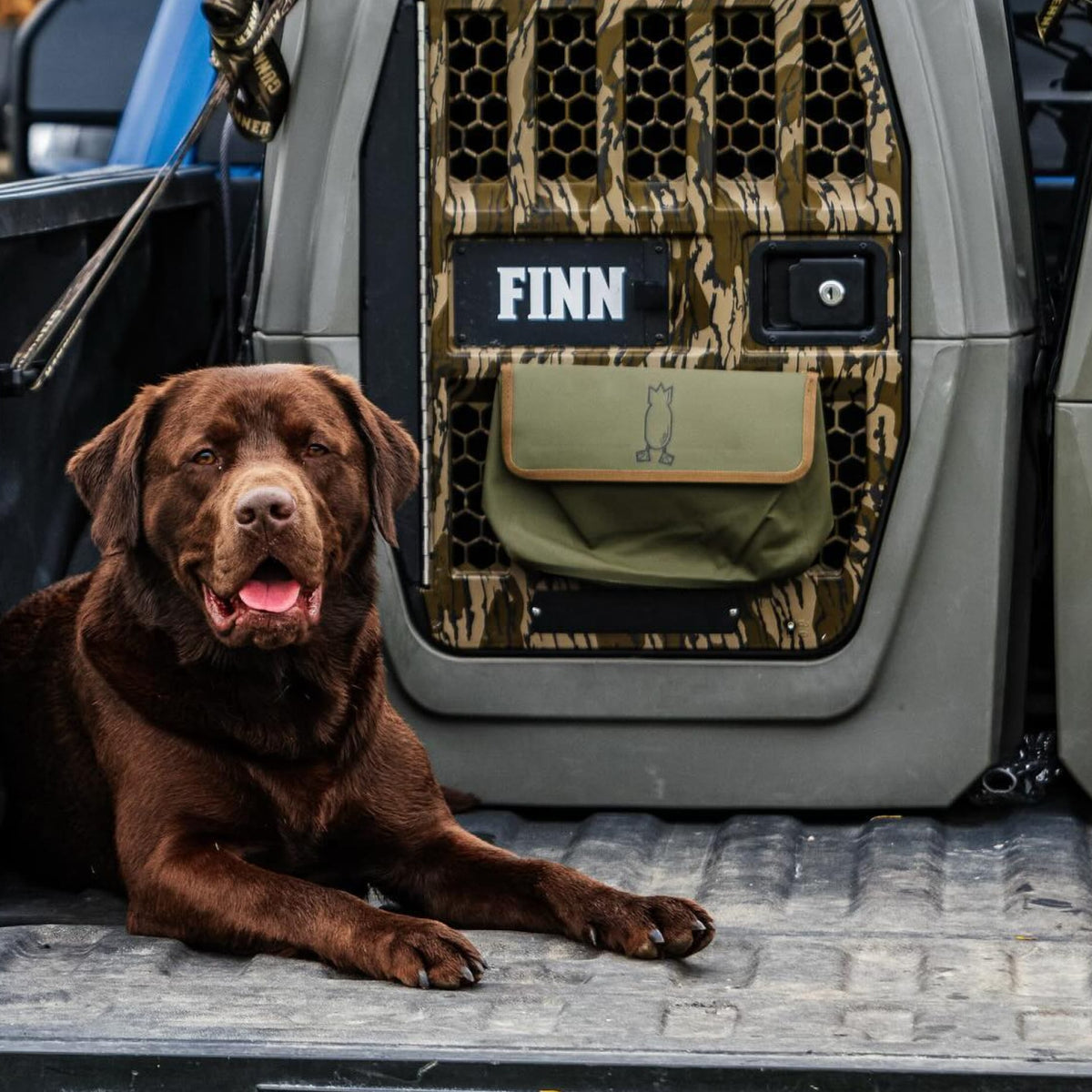 A chocolate Labrador retriever rests in the back of a vehicle beside a GUNNER G1™ KENNEL - MOSSY OAK BOTTOMLAND GUNMETAL, with a custom FINN panel and a bag featuring a simple dog illustration nearby.