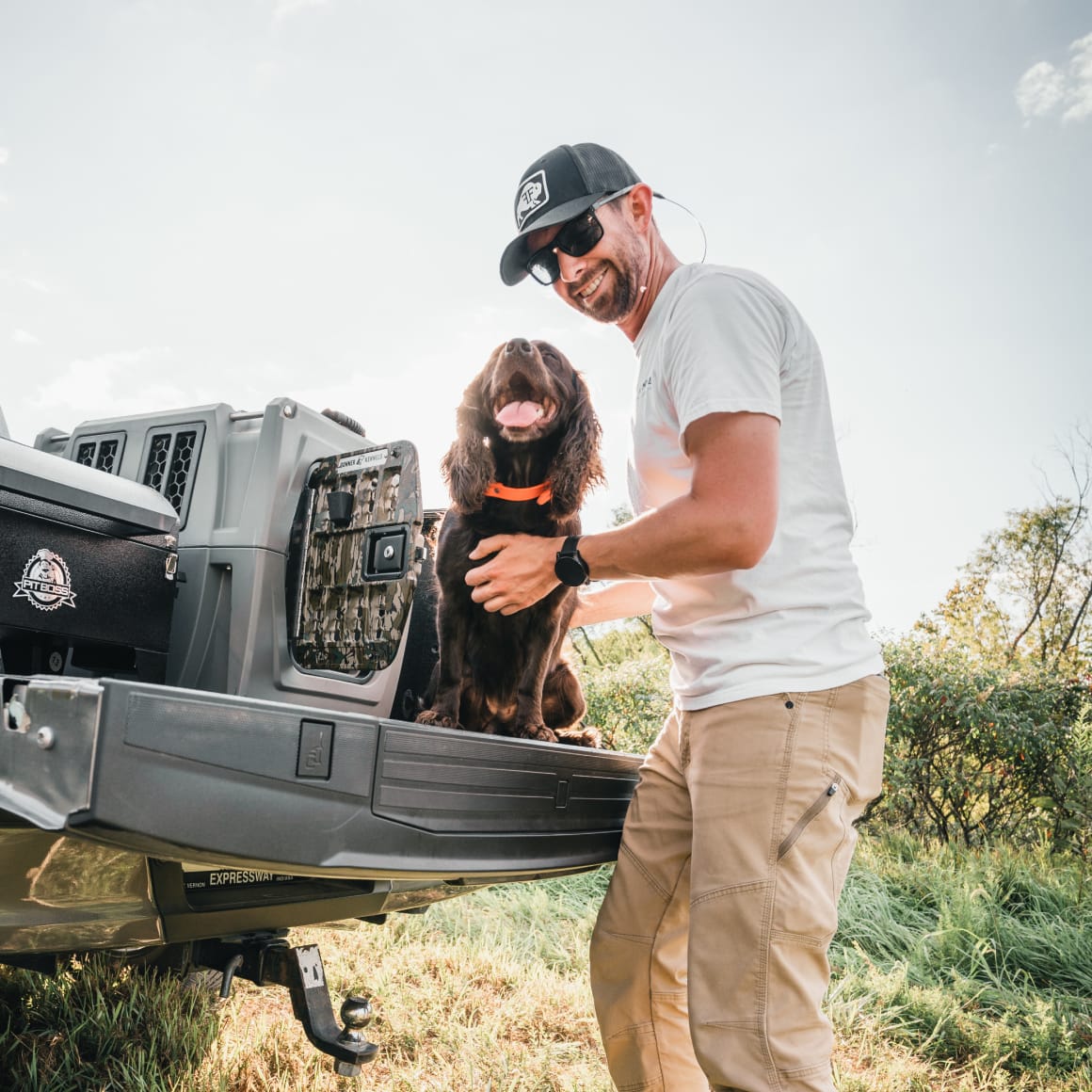 A man in sunglasses and a cap smiles beside a brown dog sitting on a truck tailgate, with the GUNNER G1™ KENNEL - MOSSY OAK BOTTOMLAND GUNMETAL crate by their side in a grassy outdoor setting.