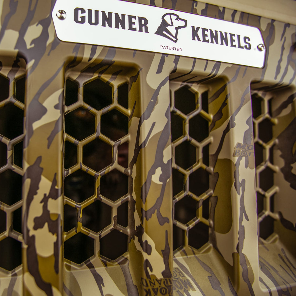 Close-up of the GUNNER G1™ KENNEL - MOSSY OAK BOTTOMLAND TAN, showing its camouflage pattern, hexagonal ventilation holes, and a metal GUNNER label at the top.