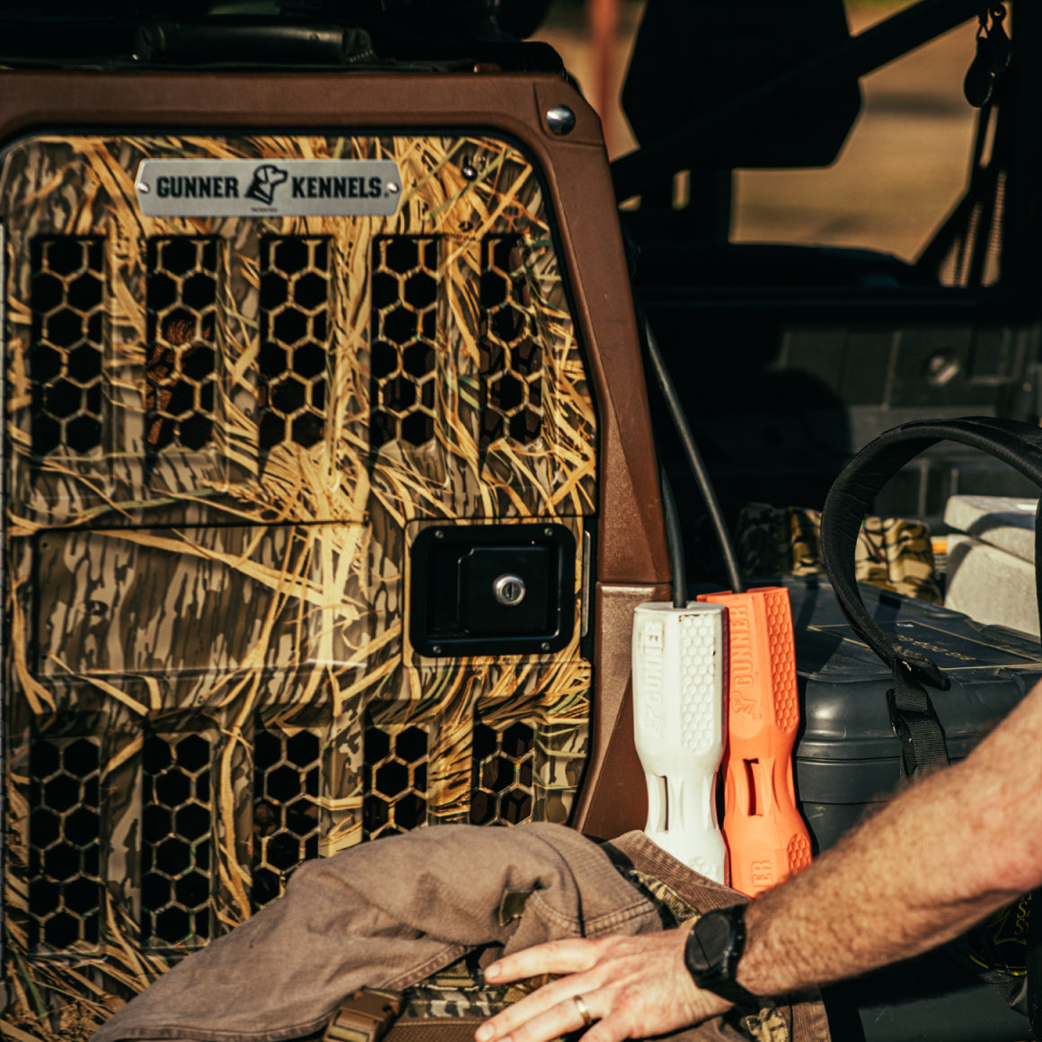 A GUNNER G1™ KENNEL in Susquehanna/Mossy Oak Shadow Grass is secured to a vehicle, with orange and white dog training dummies nearby—ideal for duck and goose hunters. A person’s arm appears in the foreground.