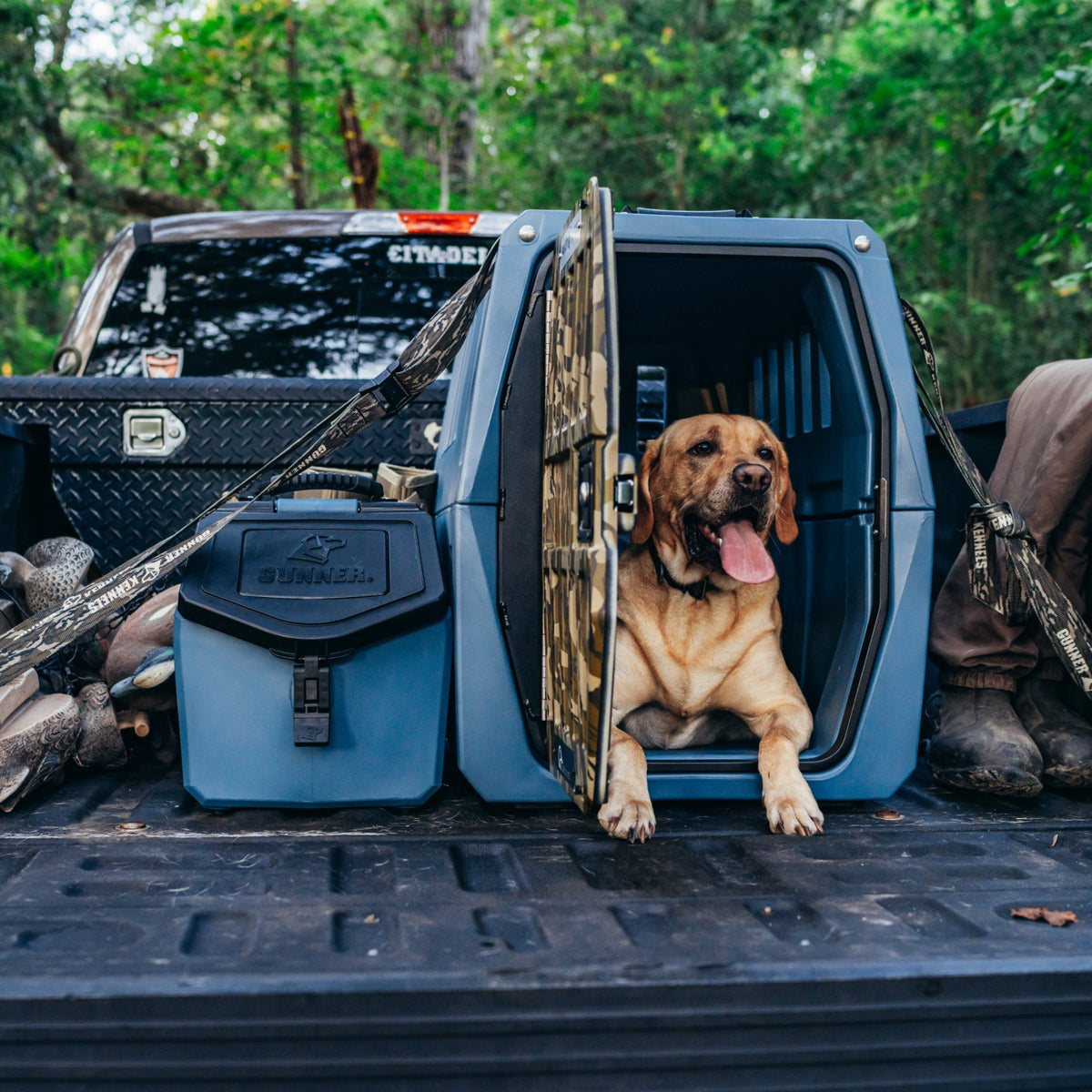 A brown dog sits in an open plastic kennel on a pickup truck bed, tongue out. The truck is parked outdoors with camping gear nearby, including a GUNNER FOOD CRATE - Ducks Unlimited Blue by GUNNER.