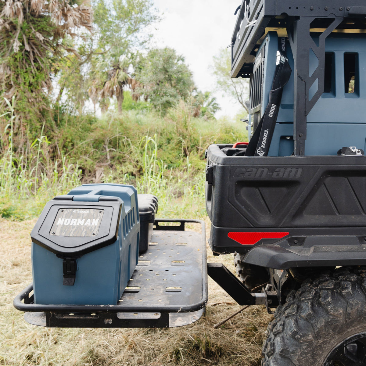 A close-up of the rear of an off-road vehicle shows GUNNER FOOD CRATE - Ducks Unlimited Blue, ideal for dog food storage, secured on a metal cargo platform; green grass and tall palm trees are visible in the background.