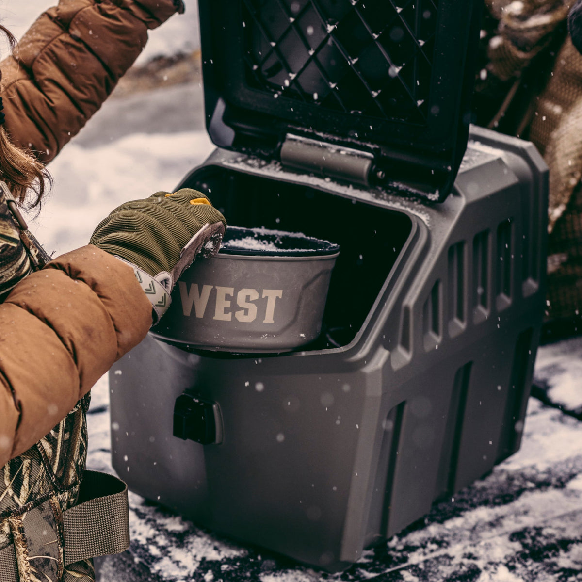 Wearing winter gear, a person retrieves the GUNNER FOOD CRATE - GUNMETAL—a pest-proof dog food container by GUNNER—from a rugged outdoor storage box on a snowy surface.