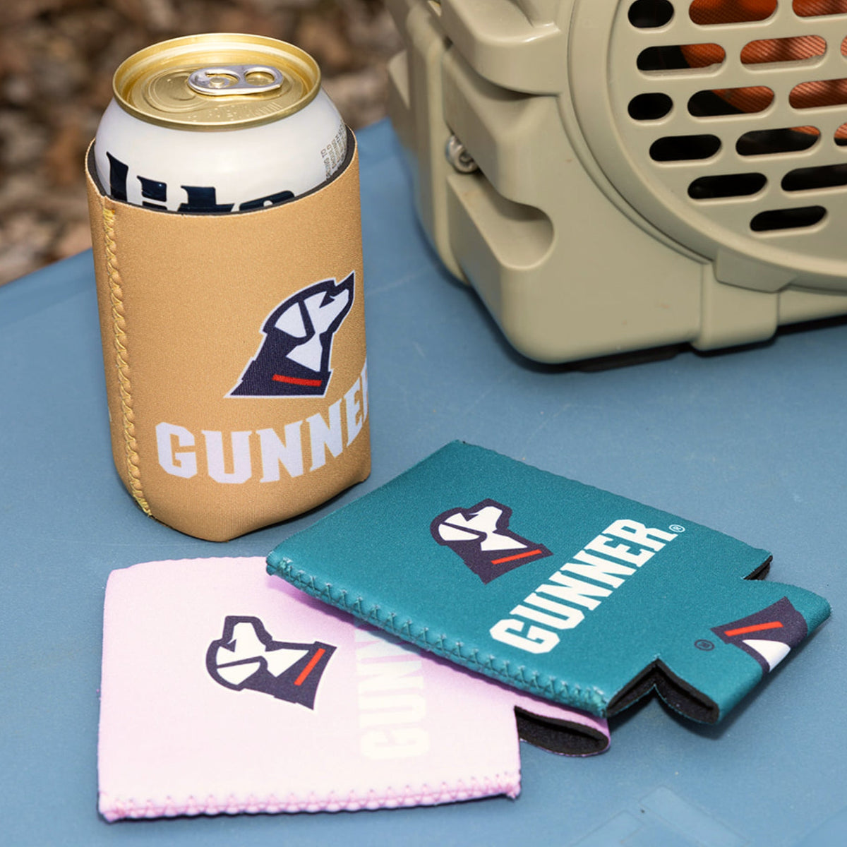 A beverage can in a tan GUNNER-branded neoprene can cooler sits beside a GUNNER KOOZIE - Pink from the Color Fetch 2025 series, with a beige ventilation device visible in the background.