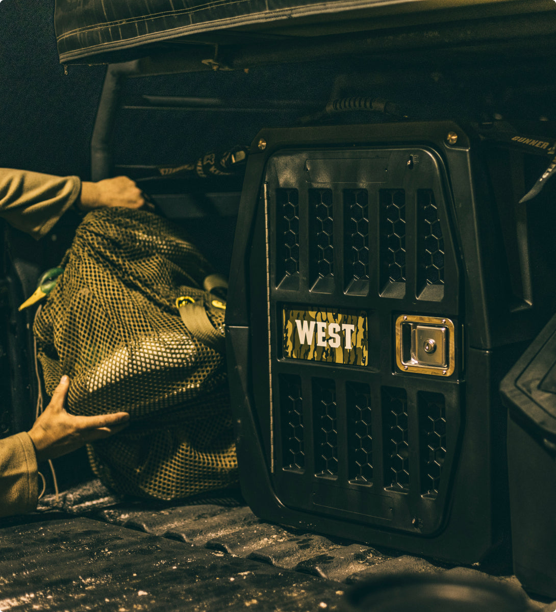 A person loads outdoor gear—a mesh bag—into the back of a vehicle next to a sturdy, black, vented crate labeled WEST with a camo-style sign, designed for a hardworking hunting dog. The area is lit with moody, yellowish light.