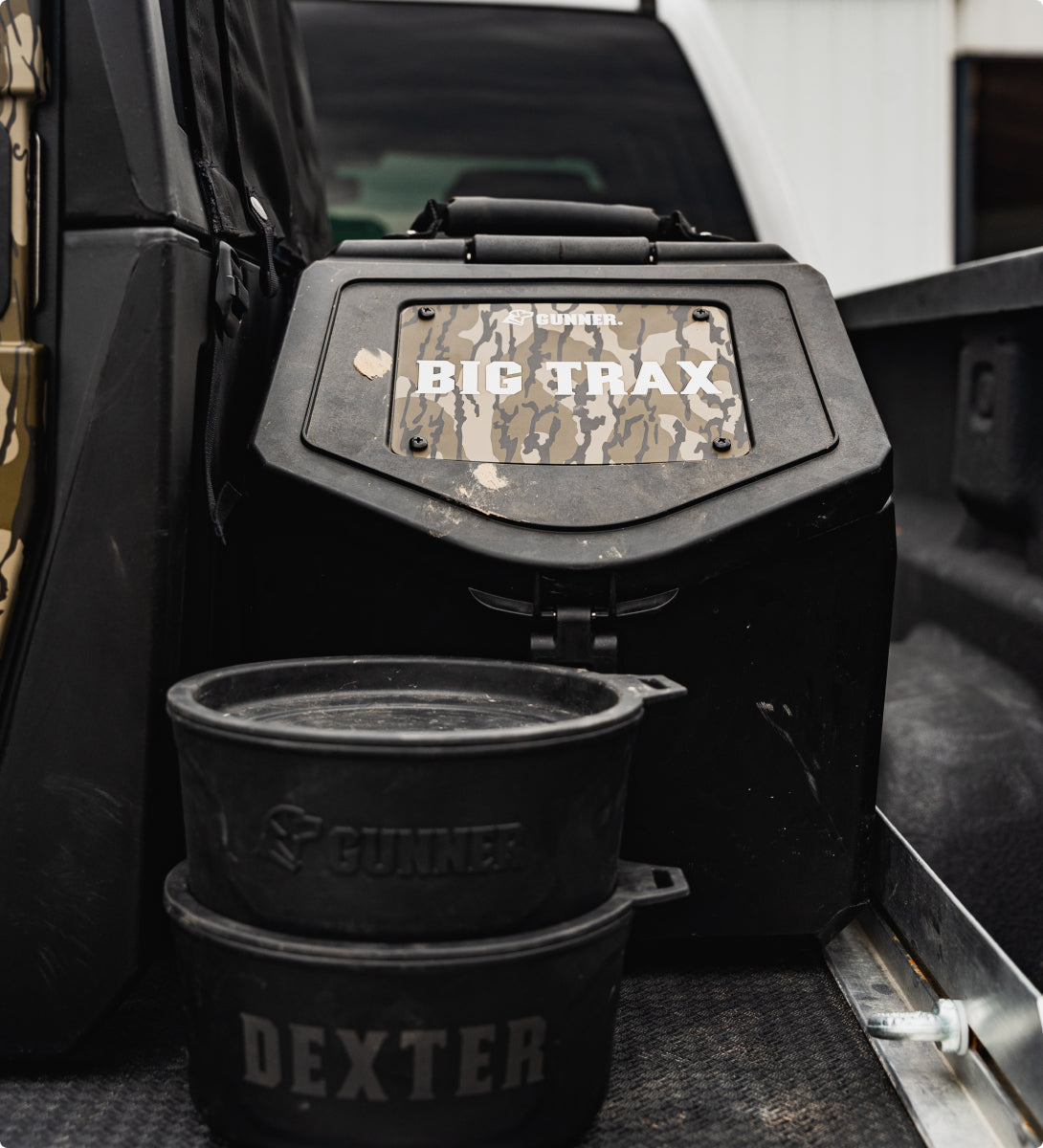 A black dog crate labeled “BIG TRAX” sits in a truck bed next to two stacked dog bowls marked “GUNNER” and “DEXTER.” The scene appears to be outdoors near a building, suggesting gear for a hunting dog.