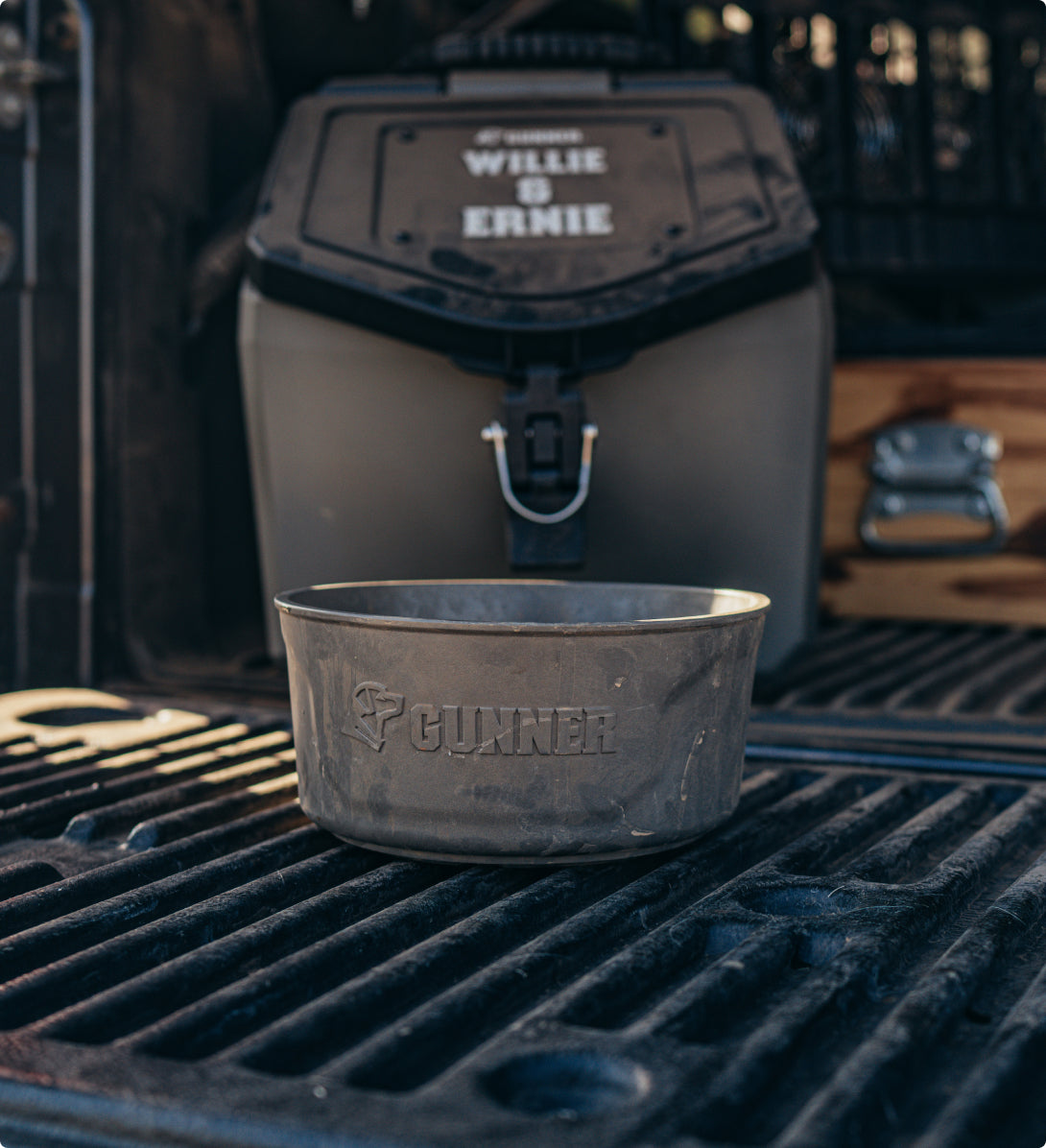 A metal bowl labeled GUNNER sits on a truck bed liner beside outdoor gear, with a large cooler labeled WILLIE & ERNIE and what appears to be a dog crate and wooden storage visible in the background.