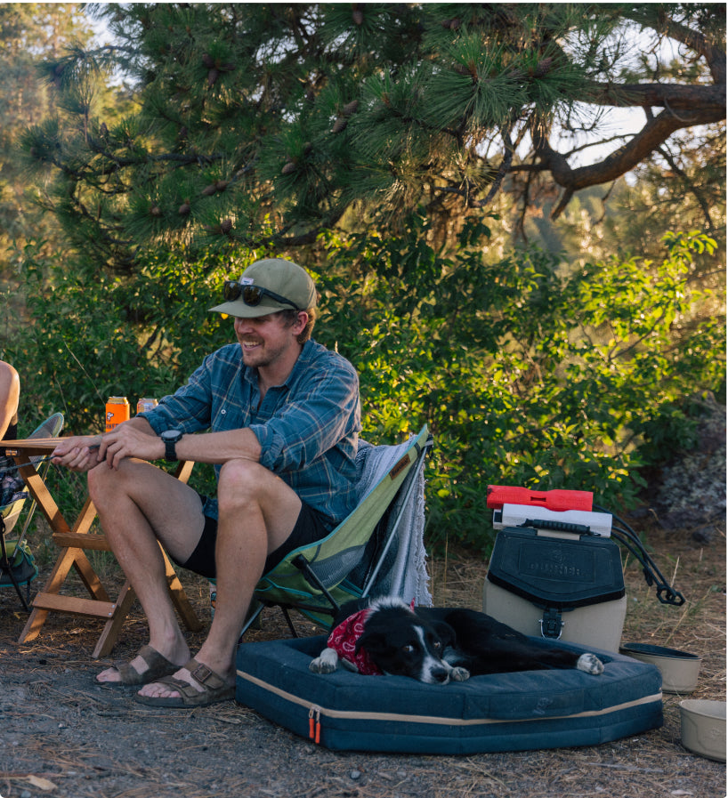 A man sits in a camping chair smiling and fishing, wearing a hat and sunglasses. A black and white working dog with a red bandana rests on a dog bed nearby. Camping gear, trees, and a portable dog crate are in the background.