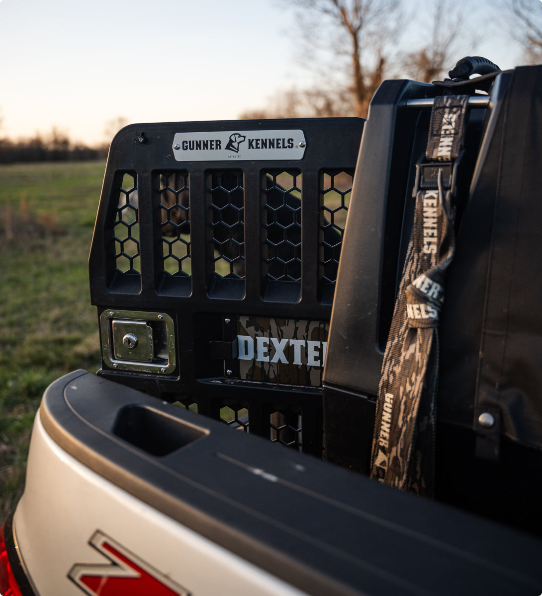 A close-up of a truck bed featuring a black Gunner Kennels dog kennel with a “DEXTER” nameplate, a camouflage strap, and part of a silver Z71 truck tailgate—perfect for transporting your hunting dog—outdoors at sunset.
