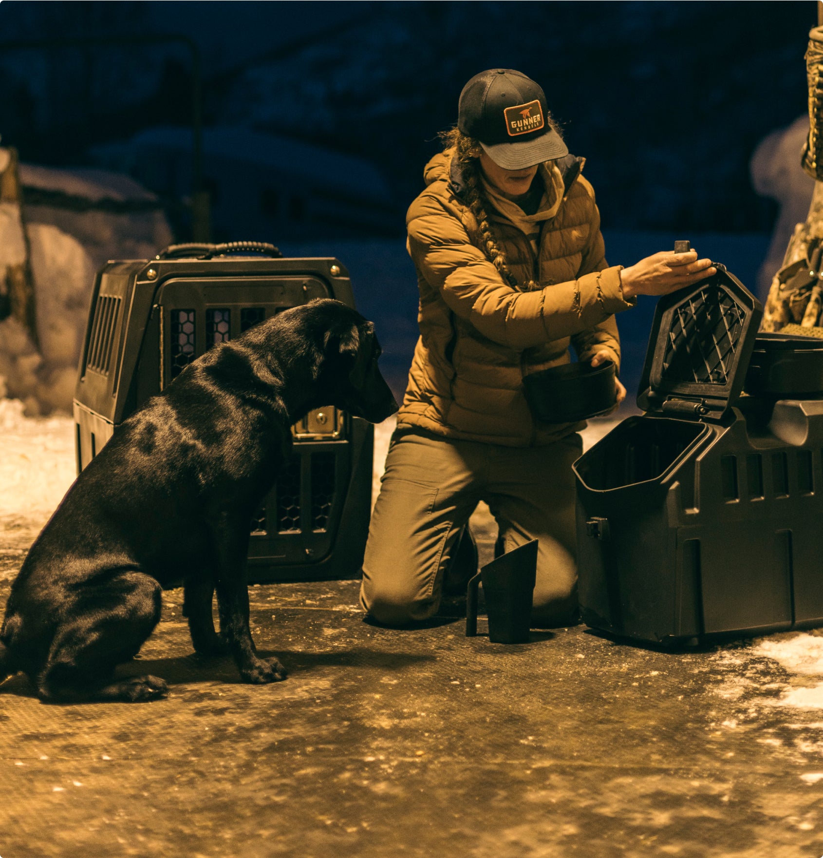 A person in a jacket and hat kneels on snowy ground beside a black hunting dog, opening a black plastic container. Two large dog crates are nearby, and it appears to be evening.