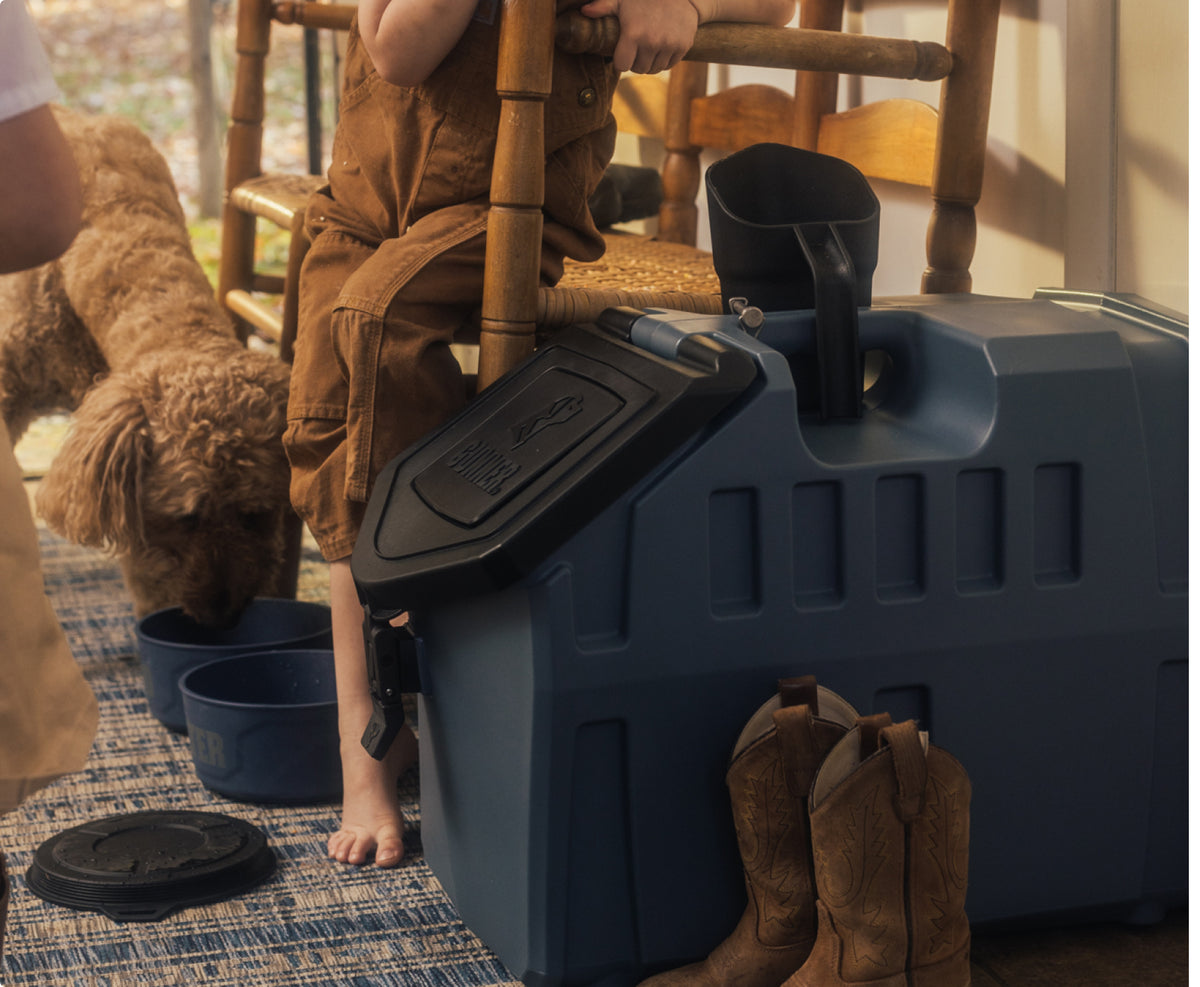 A brown dog drinks from a blue bowl on the floor beside a large gray dog kennel. Two children, one barefoot, sit nearby with cowboy boots and outdoor gear on the floor. The scene is indoors with wooden furniture visible.