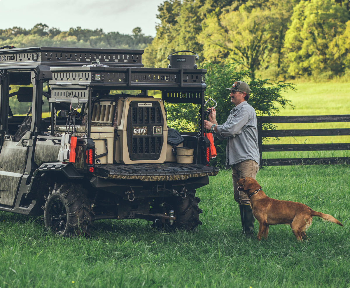 A man stands by an off-road vehicle in a grassy field, preparing gear with his working dog beside him. The vehicle holds a dog crate and outdoor equipment. Trees and a wooden fence are in the background.