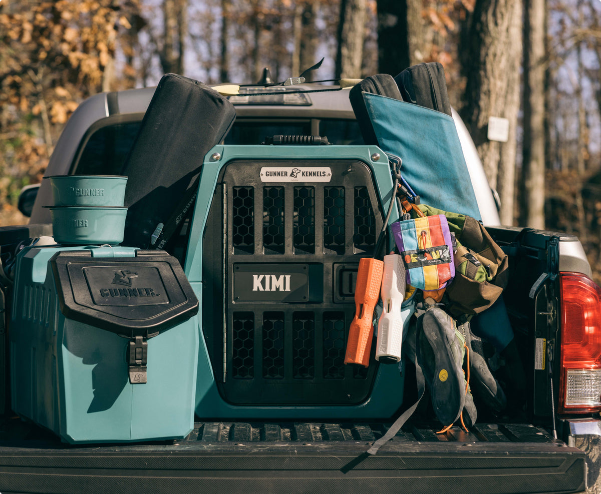 A pickup truck bed loaded with outdoor gear, including a large teal dog crate labeled Kimi for a hunting dog, food containers, water bowls, bags, and dog toys, surrounded by trees in a wooded area.
